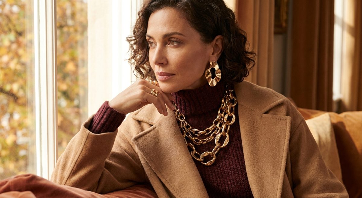Femme en manteau camel portant un collier chunky à maillons dorés et des boucles d'oreilles sculpturales, tendances automne-hiver 2026