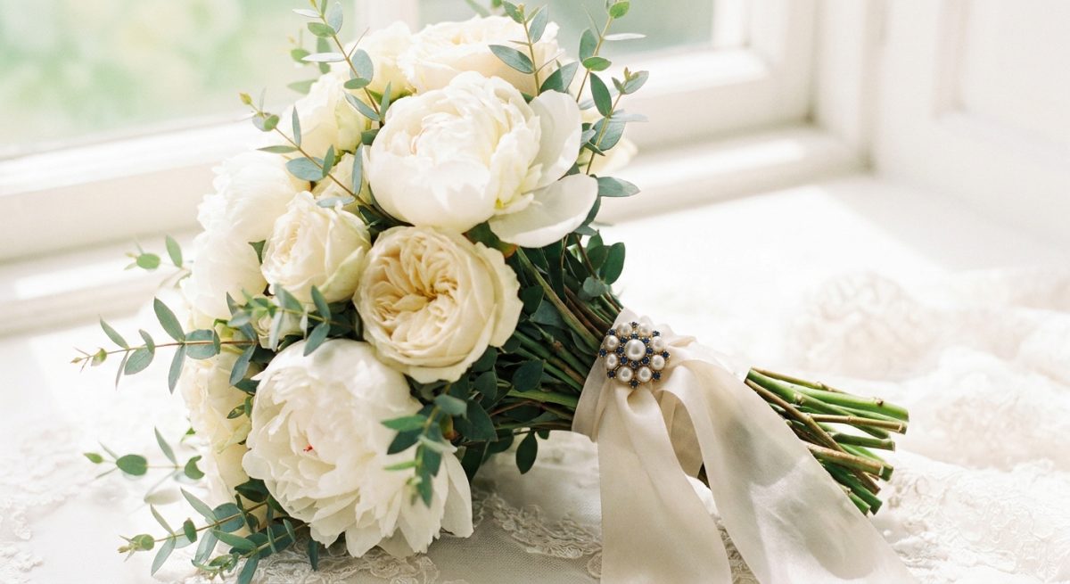 Bouquet de mariée en pivoines blanches et roses crème avec eucalyptus, ruban de soie crème, et broche perle et saphir épinglée à la base du bouquet