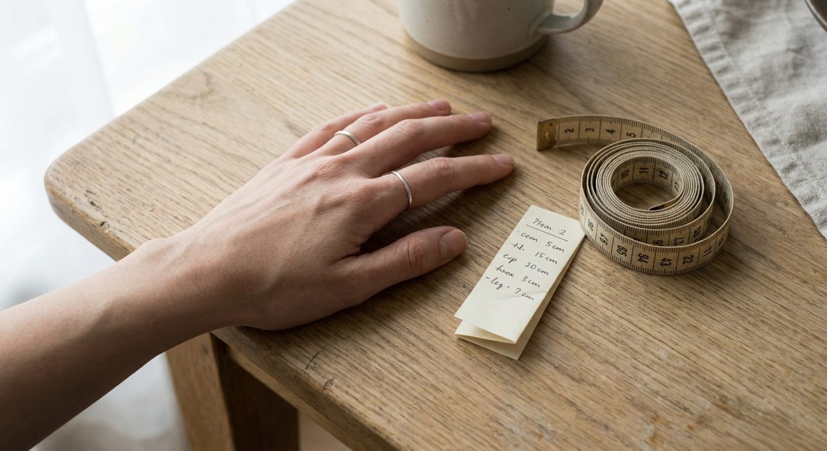 Main féminine posée sur une table en bois clair avec un mètre ruban et un petit papier de mesure