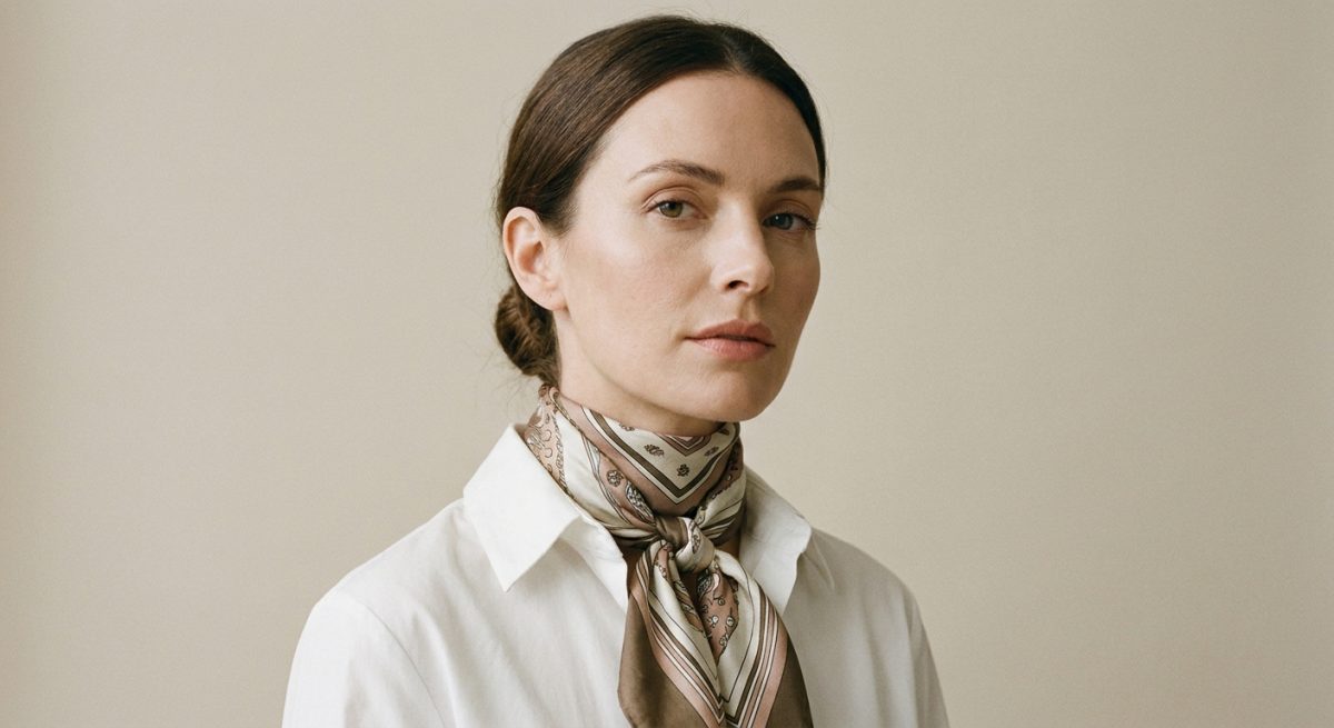 Femme trentenaire portant un foulard carré soie noué en ascot sous un col de chemise blanche, portrait trois-quart sur fond beige clair