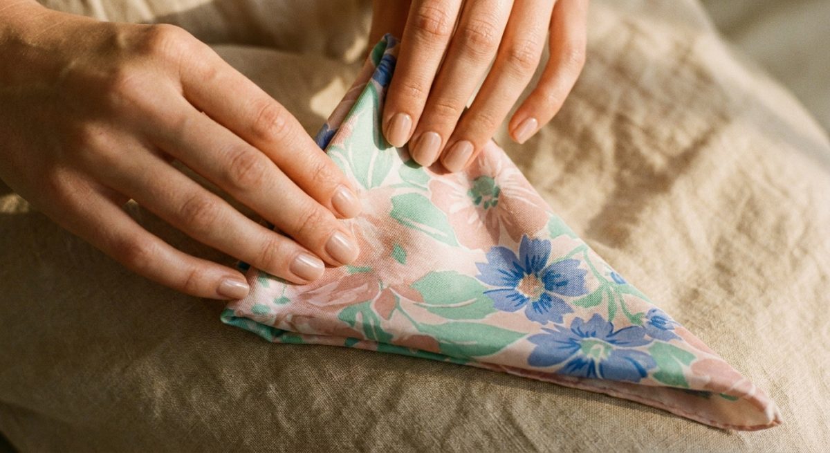 Mains pliant un carré de soie pastel en triangle sur lin beige, lumière dorée d'après-midi